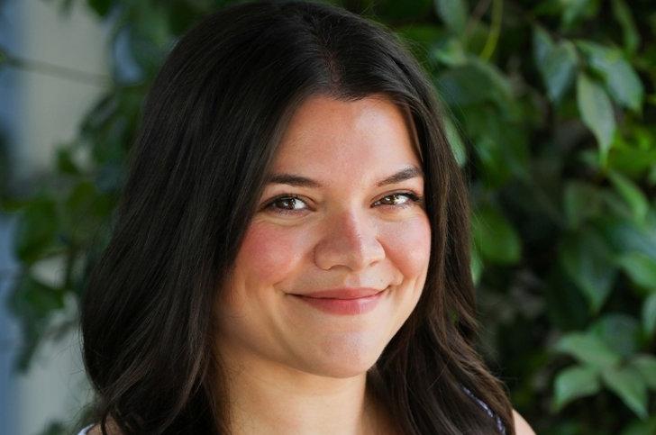 Close-up headshot of Emily Butts, who has long wavy dark hair and stands in front of a green ivy wall.